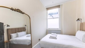 A hotel room with two beds, a large window, a radiator, and a mirror with an ornate frame. - Elegant South Kensington Retreat (London)