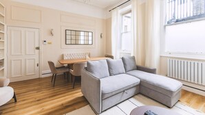 A modern living room with a grey sectional sofa, a wooden dining table with chairs, a chandelier, and a large window with curtains. - Elegant South Kensington Retreat (London)