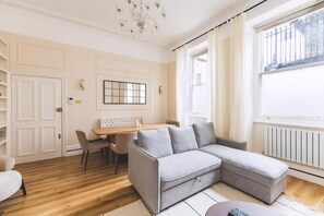 A modern living room with a grey sectional sofa, a wooden dining table with chairs, a chandelier, and a large window with curtains. - Elegant South Kensington Retreat (London)