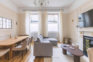 A living room with a sofa, a coffee table, a TV, a fireplace, and a dining area with chairs and a table. - Elegant South Kensington Retreat (London)