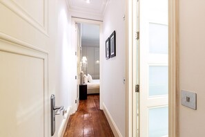 A hallway with a wooden floor, white walls, and a door leading to a bedroom with a bed and a nightstand. - Boutique Kensington Retreat Near Museums (London)