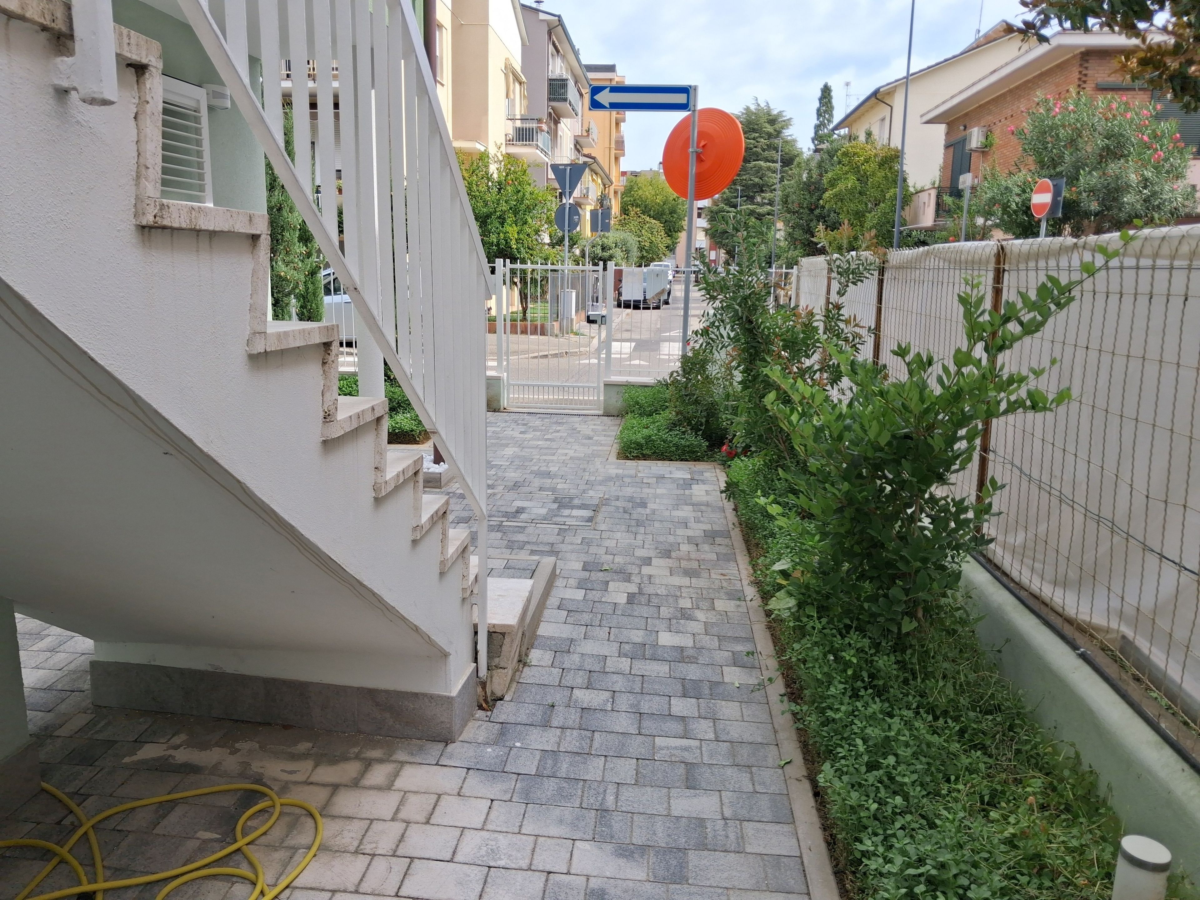 Un étroit sentier pavé mène à une entrée fermée par un portail, de part et d’autre de laquelle se trouvent un bâtiment blanc et une clôture verte.