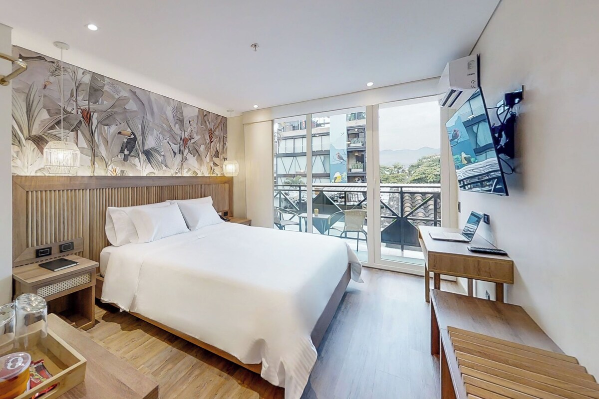 A modern hotel room with a large bed, a wooden headboard, a balcony with a view, a desk with a laptop, and a wall-mounted TV.