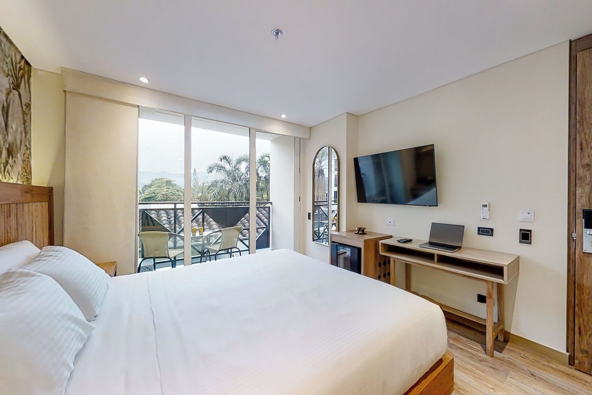A hotel room with a bed, a desk with a laptop, a TV, and a balcony with outdoor furniture.