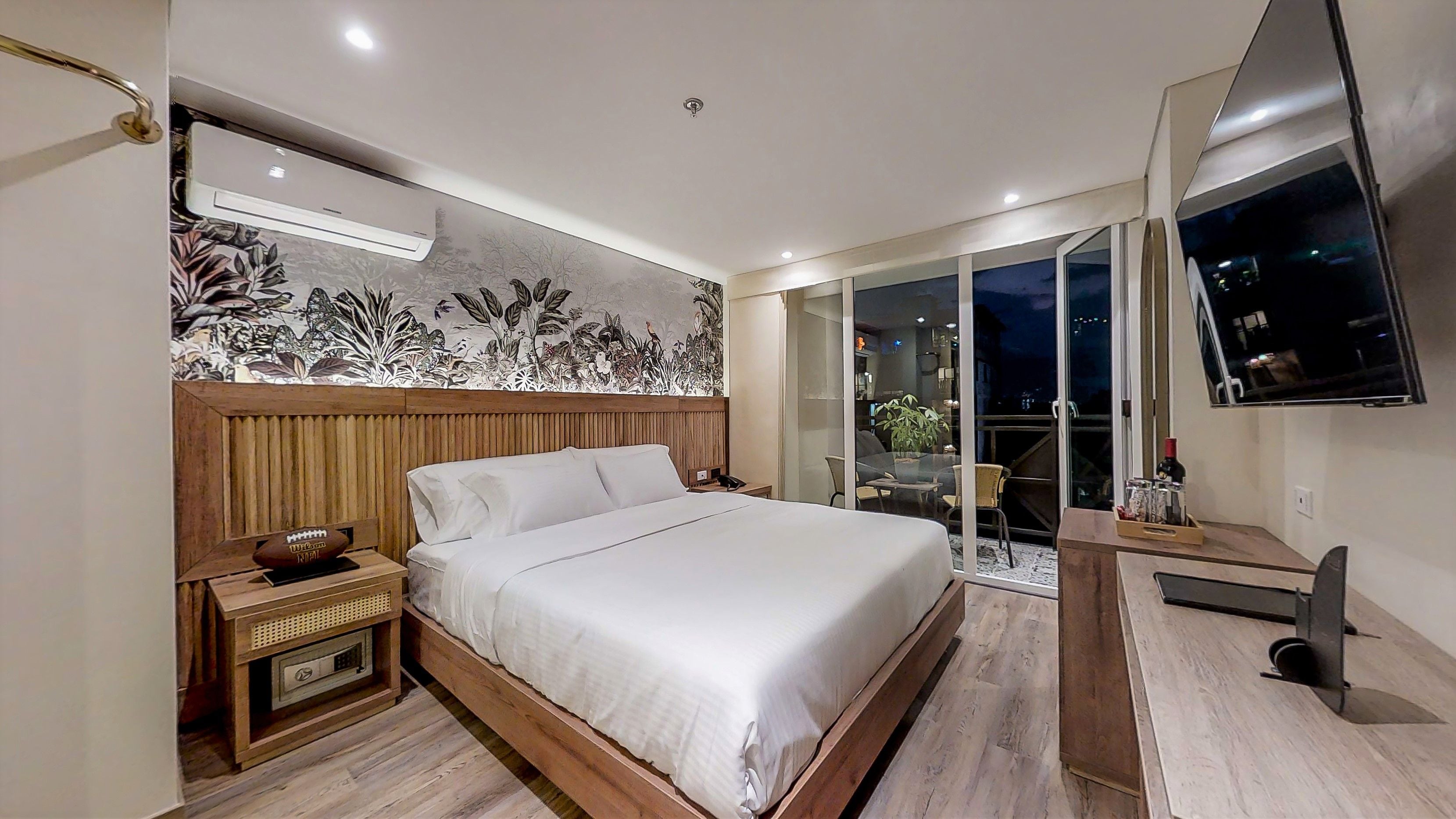 A modern hotel room with a large bed, a wooden headboard, a flat-screen TV, a desk with a chair, and a balcony with a view of the city at night.