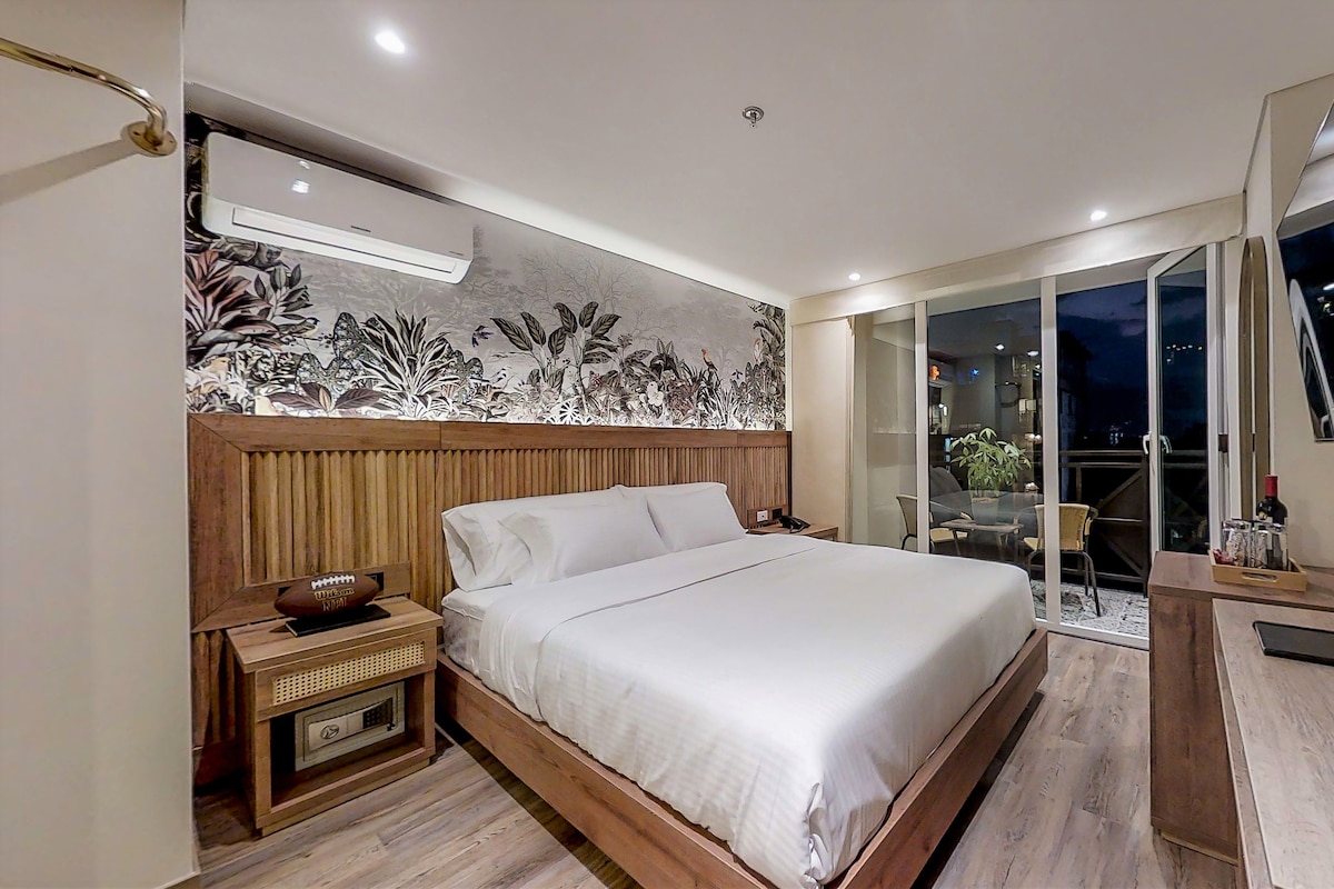 A modern hotel room with a large bed, a wooden nightstand, a desk with a chair, a flat-screen TV, and a balcony with a view.