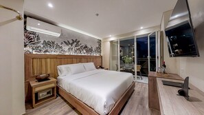 Una habitación de hotel moderna con una cama grande, cabecera de madera, televisión de pantalla plana, escritorio con silla y balcón con vista a la ciudad de noche.