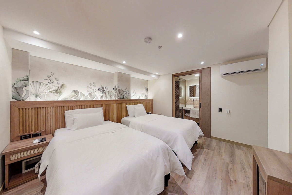 A hotel room with two beds, a wooden headboard, a wall-mounted air conditioner, and a bathroom visible through an open door.