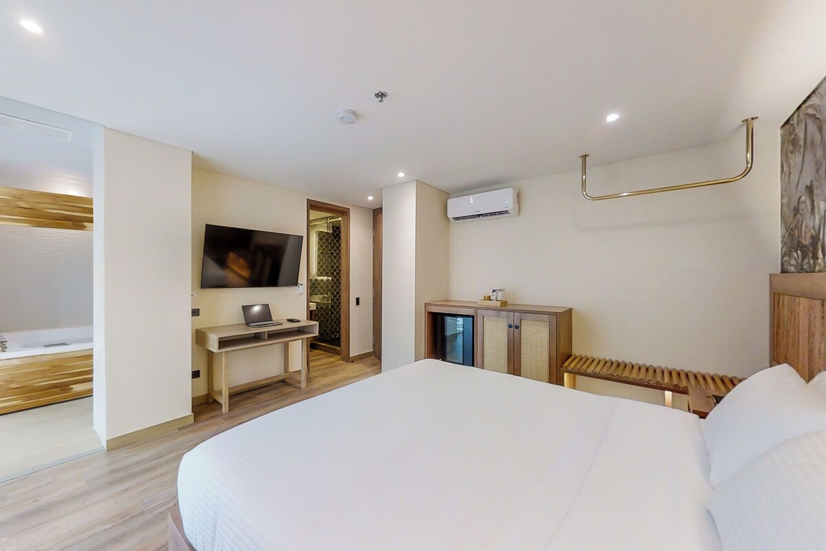 A modern hotel room with a large bed, a desk with a laptop, a television, and a bathroom with a shower.