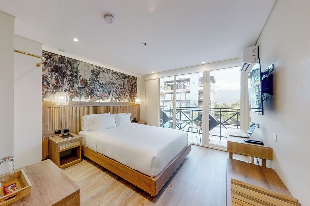 A modern hotel room with a large bed, a wooden headboard, a balcony with a view, and a flat-screen TV mounted on the wall.