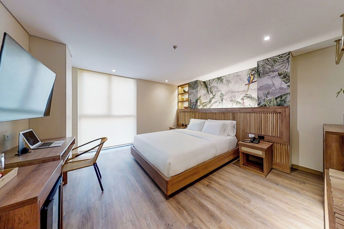 A modern hotel room with a large bed, a desk with a laptop, a flat-screen TV, and a wooden floor.