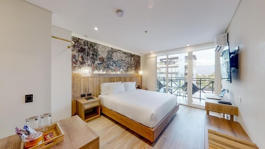Una habitación de hotel moderna con una cama grande, cabecera de madera, mesitas de noche, un escritorio y un balcón con vistas.