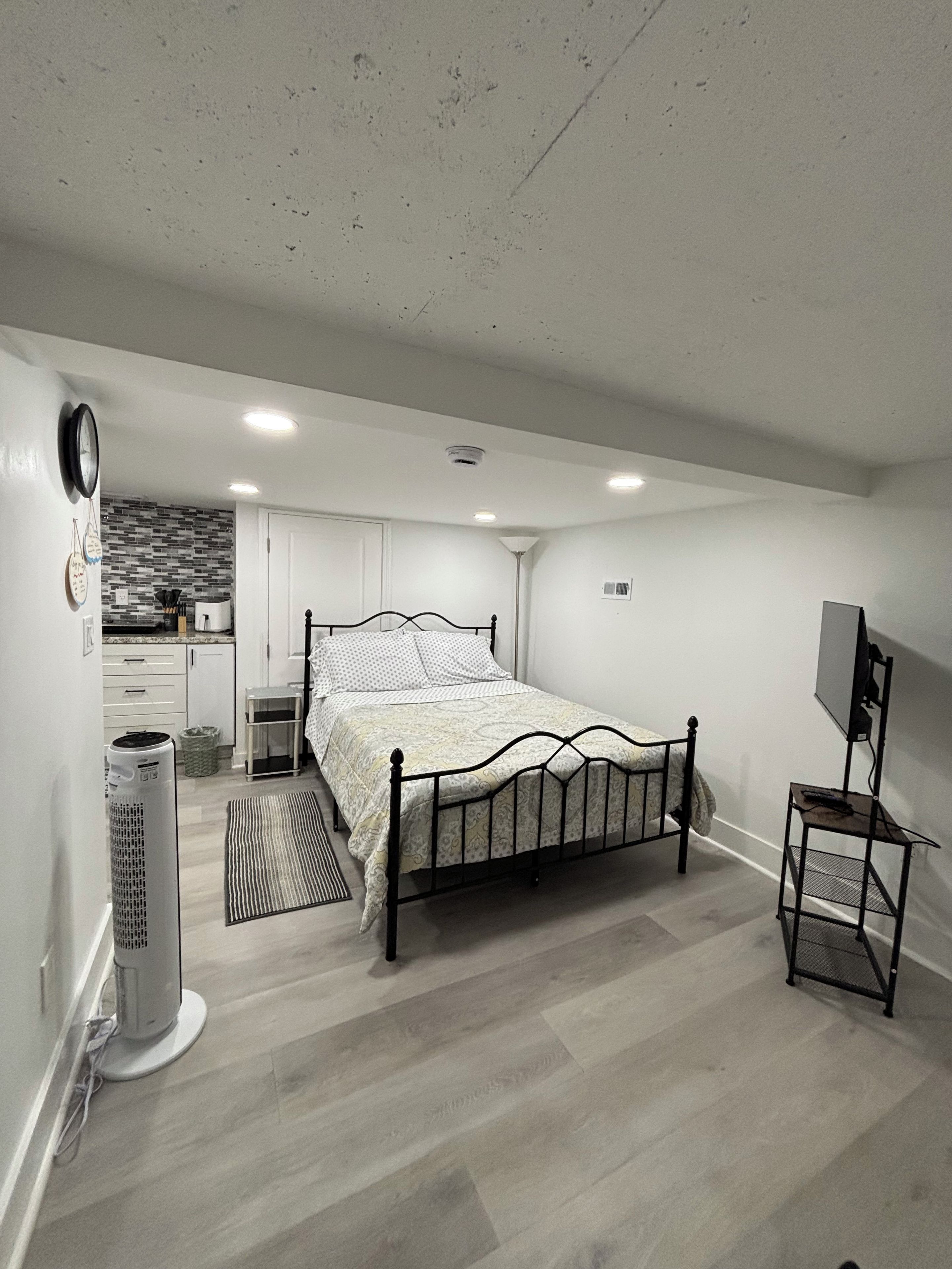 Un dormitorio con una cama, un calentador, un mueble para televisión y una kitchenette al fondo.