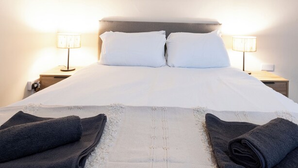 A neatly made bed with white linens and two pillows, flanked by two bedside lamps on wooden nightstands. - Bayview Aparthotel by Guestz (Morecambe)
