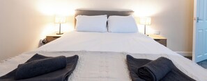 Una cama bien hecha con sábanas blancas y dos almohadas, flanqueada por dos lámparas de noche sobre mesitas de madera.