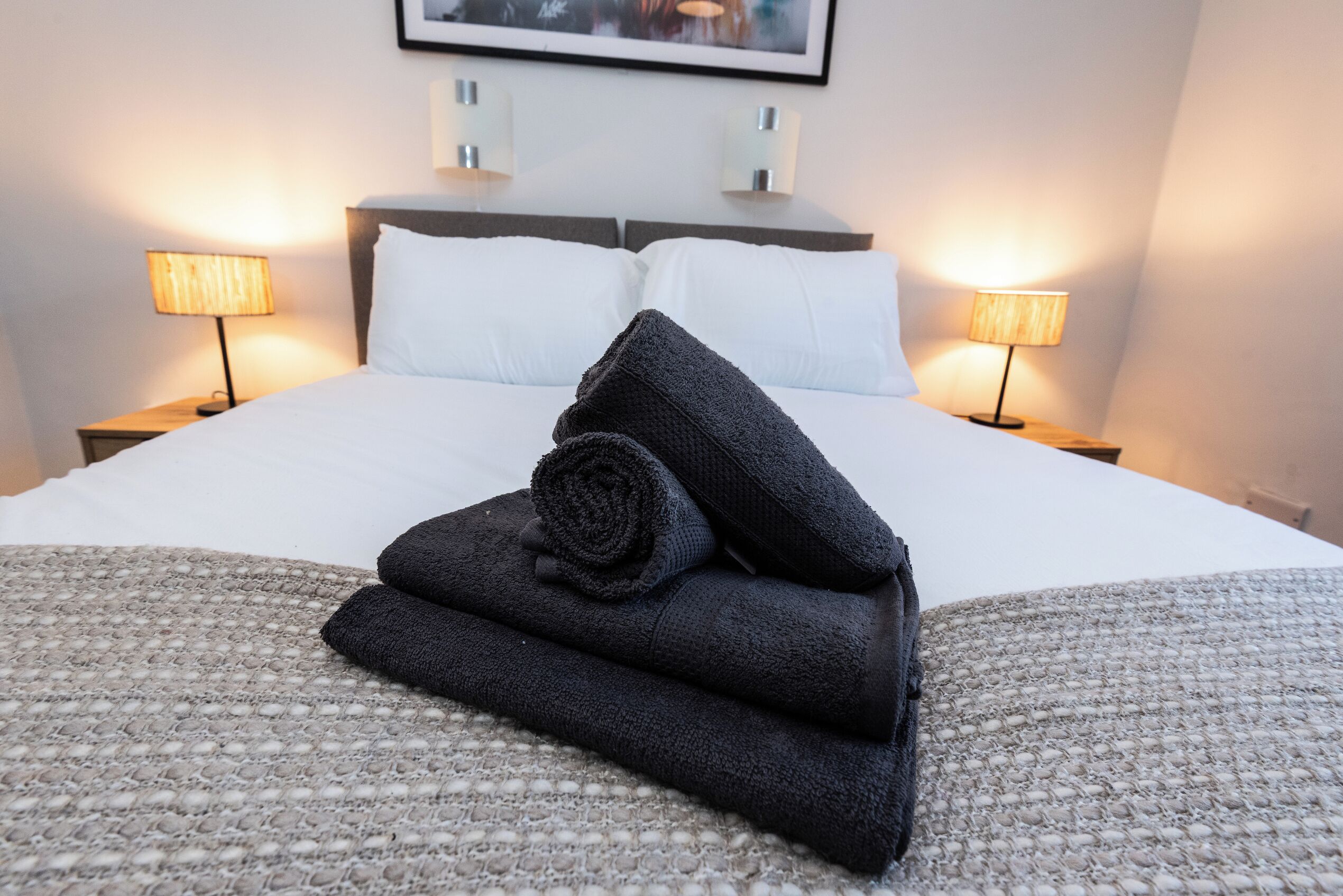 Een netjes opgemaakt bed met opgerolde handdoeken, een hoofdbord, twee bedlampen en een ingelijste foto aan de muur.