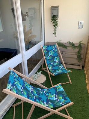 Dues cadires de saló de fusta amb tapisseria floral, una porta de vidre, una petita banca de fusta i un planter penjant a la paret.