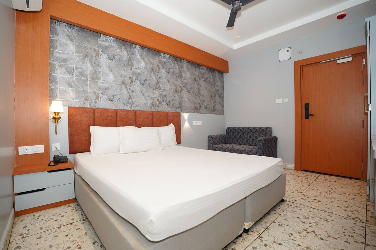 Una habitación de hotel con una cama grande, una silla, cabecera de madera, una lámpara de pared y una puerta.