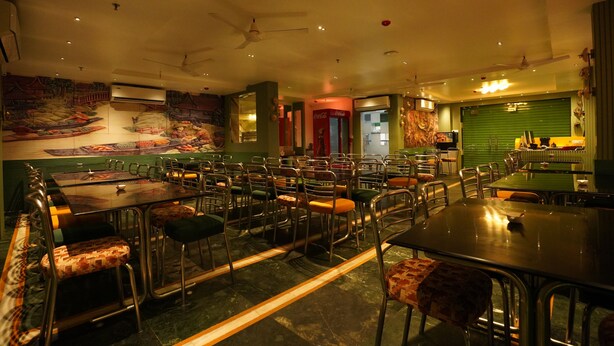 A restaurant with multiple tables and chairs, ceiling fans, and a green wall with a mural. - Bidesh Ghar Hotel (Puri)