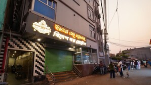 A hotel with a neon sign, a green shuttered entrance, and a staircase leading to the entrance. - Bidesh Ghar Hotel (Puri)