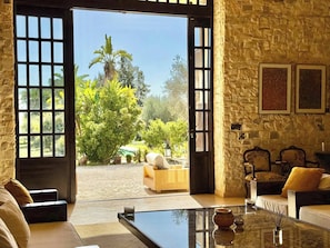 A room with a stone wall, a glass coffee table, and a view of a garden with a palm tree. - Artist's House Villa in Utique (Utica)