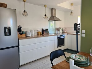 Eine moderne Küche mit Kühlschrank, Herd und Kaffeemaschine. Hängelampen und ein Fenster sorgen für natürliches Licht.