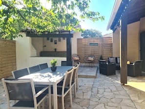 Une terrasse extérieure avec un sol en pierre, une table avec des chaises, un fauteuil suspendu, une petite plante en pot et une clôture en bois.