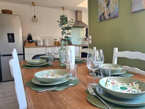 Ein Esszimmer mit Tisch, gedeckt mit grünen Tellern, Schüsseln und Weingläsern, eine Küche im Hintergrund und eine grüne Wand mit einem Gemälde.