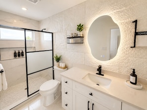 A modern bathroom with a glass shower enclosure, a white toilet, a vanity with a large mirror, and a towel rack. - Muse Atelier | Houston (Houston)