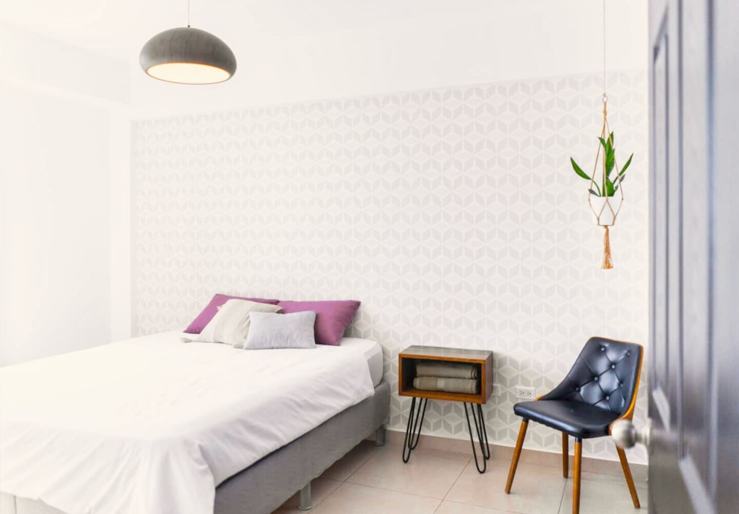 A bedroom with a bed, a chair, a side table, and a hanging plant.