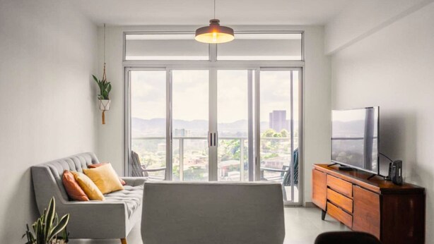 A modern living room with a sofa, a TV, a wooden cabinet, and a large window offering a city view. - Nice City View Apartment in Escalon! (San Salvador)