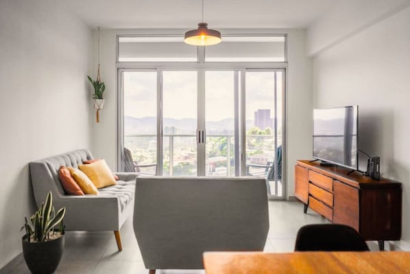 Un salon moderne comprenant un canapé, un téléviseur, un meuble en bois et une grande fenêtre donnant sur la ville.