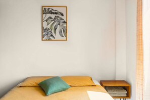 A bedroom with a bed, a green pillow, a wooden nightstand, and a framed plant artwork on the wall.