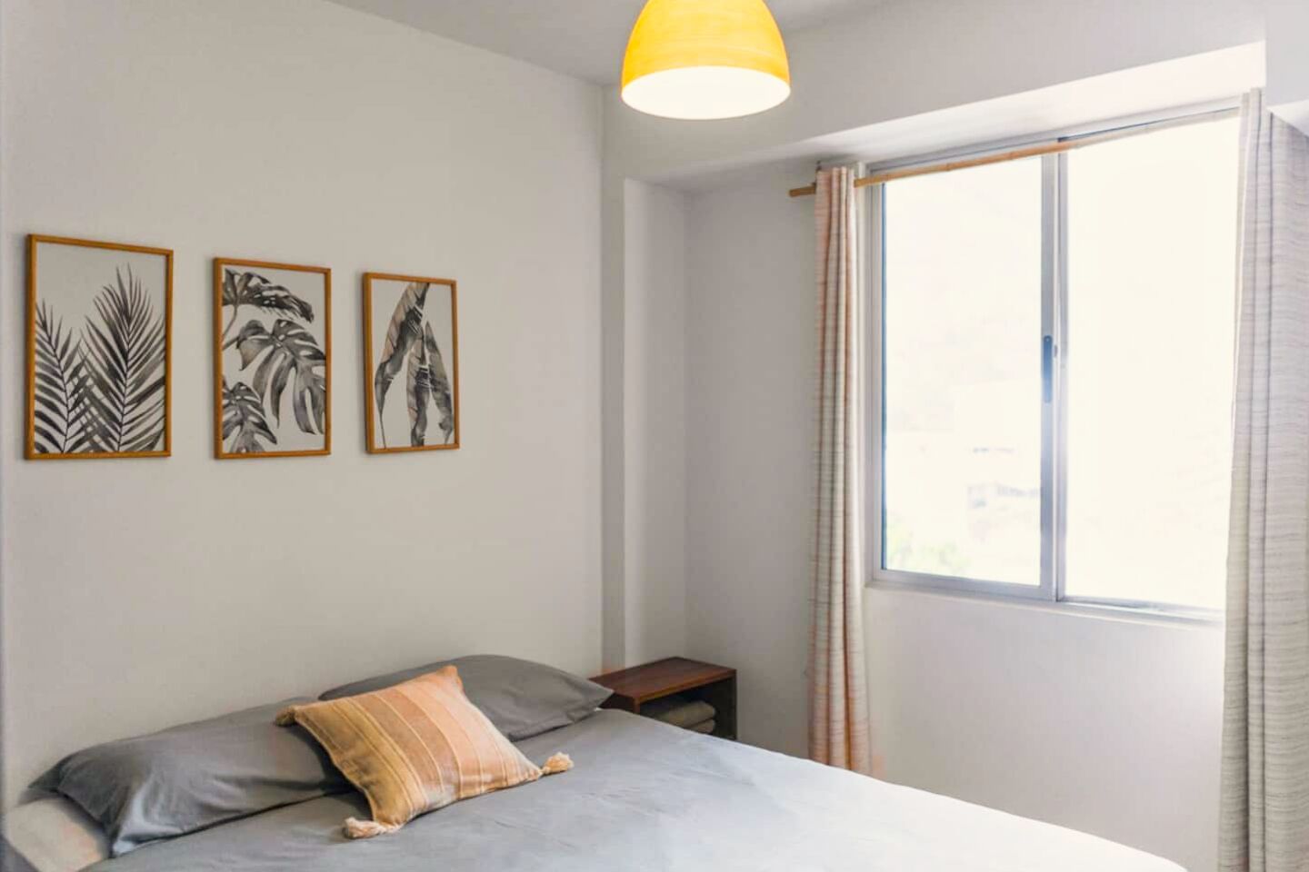 Un dormitorio con cama, mesita de noche y ventana con cortinas. Se observan tres cuadros enmarcados en la pared.