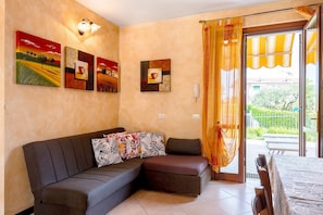A living room with a sofa, a table, and a view of greenery outside. - Gigli Apartment with Garden, Shared Pool, Wi-Fi and Air Conditioning (Lazise)
