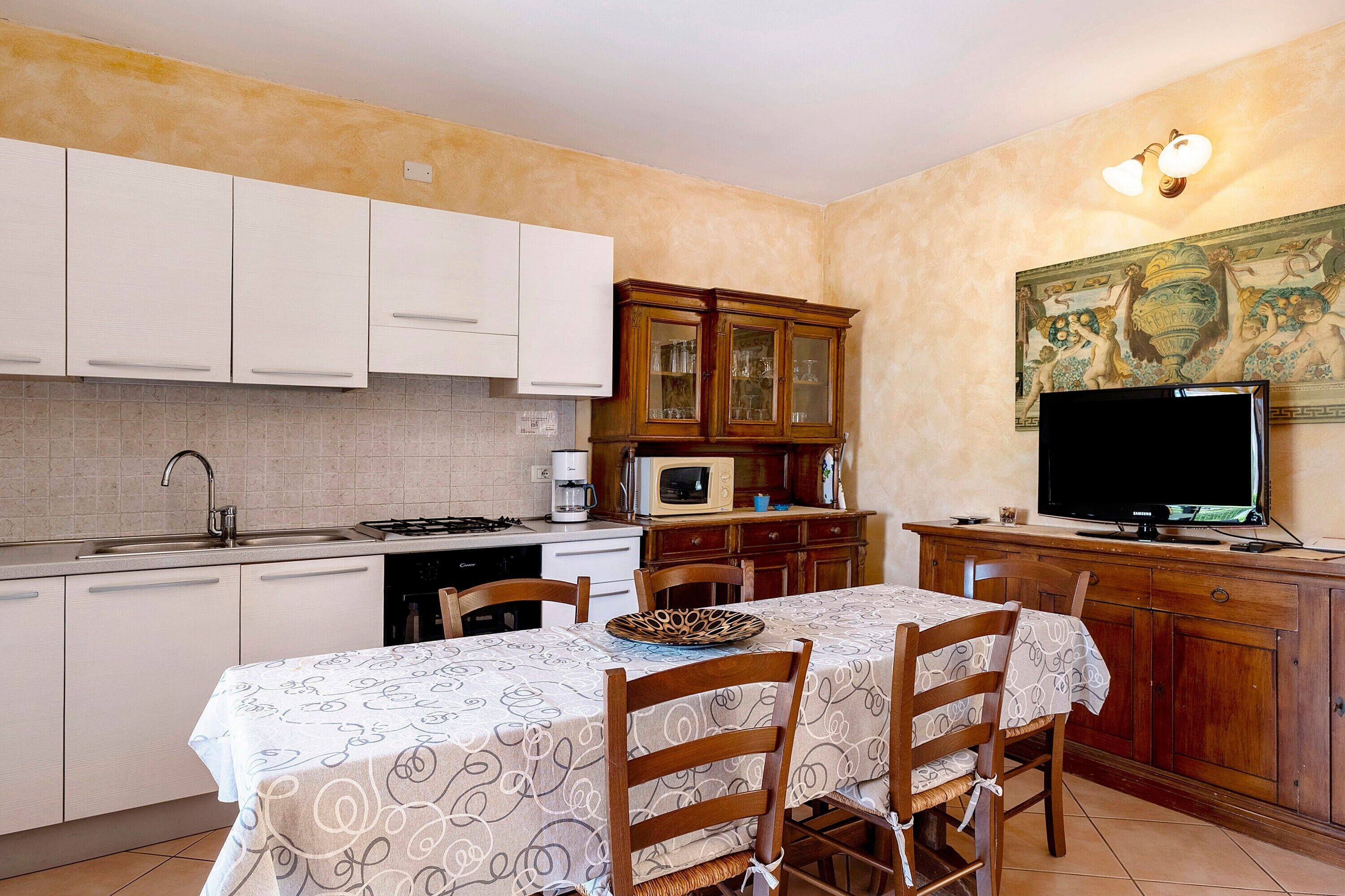 Une cuisine compacte avec des meubles blancs, une table à manger avec des chaises, un four à micro-ondes et un téléviseur.