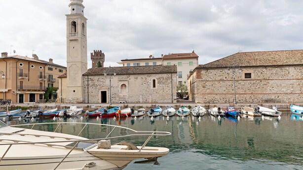 Una marina amb vaixells amarrats, un edifici històric amb una torre, i una paret de pedra.