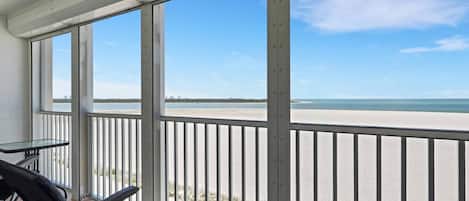 A balcony with a view of the beach and ocean, a table, and two chairs.