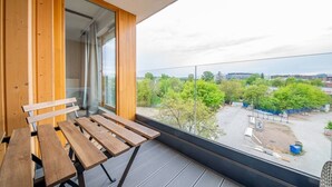 Een balkon met een houten bankje en tafel, een glazen deur en uitzicht op een parkeerplaats met bomen en gebouwen.
