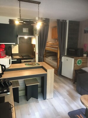Eine kompakte Küche mit Mikrowelle, Kaffeemaschine und kleinem Kühlschrank. Ein Holztisch mit Hocker, ein Etagenbett und ein kleiner Fernseher sind ebenfalls vorhanden.