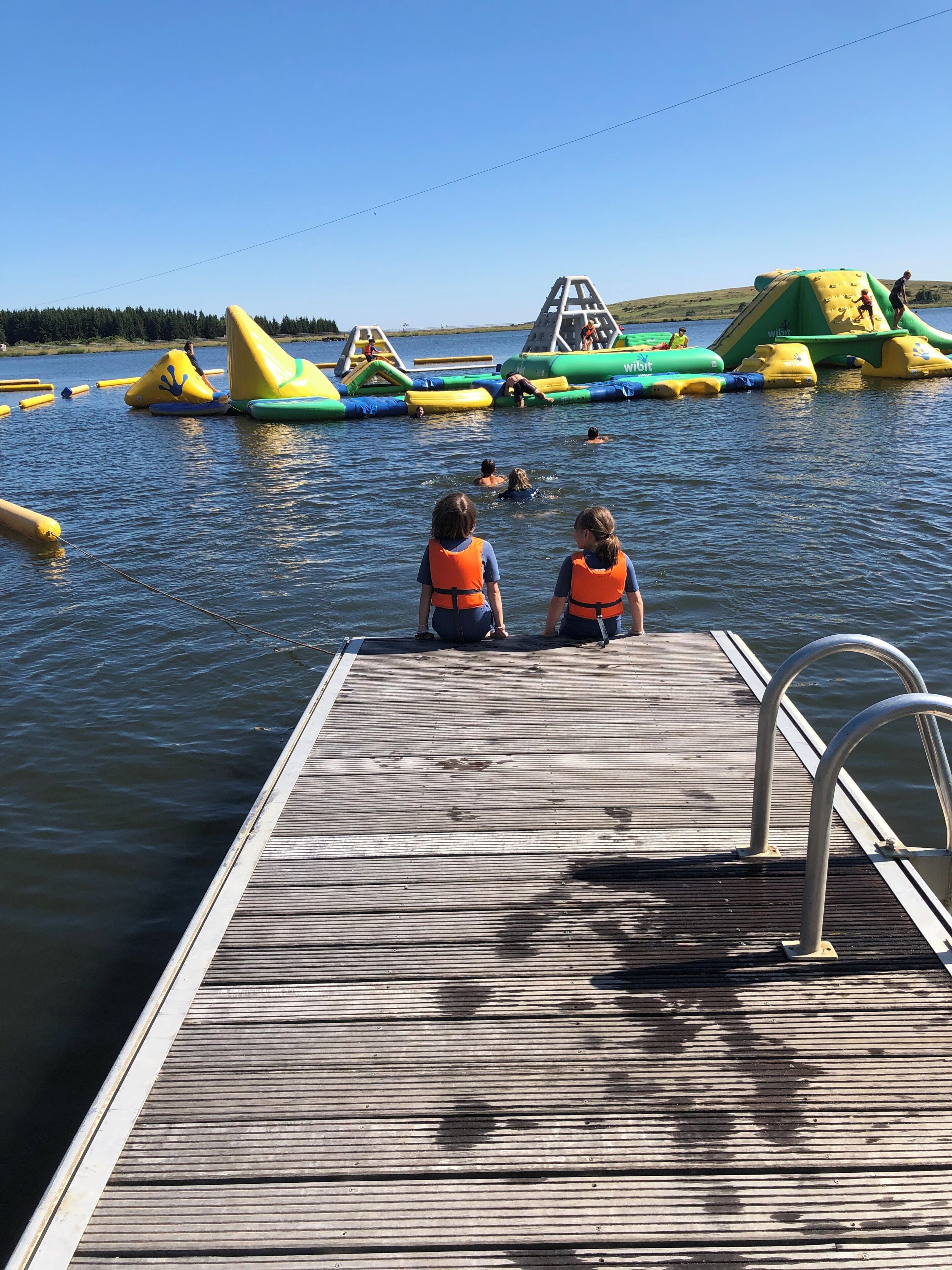 Une jetée en bois s’étendant sur une étendue d’eau, avec des structures gonflables pour jeux aquatiques en arrière-plan.