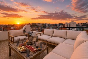 Una terraza en la azotea con sofá, mesa de comedor y sillas, con vistas a un paisaje urbano al atardecer.