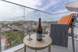 A bottle of wine on a balcony table with two glasses, a view of a cityscape, and a chair with a blanket. - Premium Lisbon Residence | Pool, Gym & Parking (Lisboa)