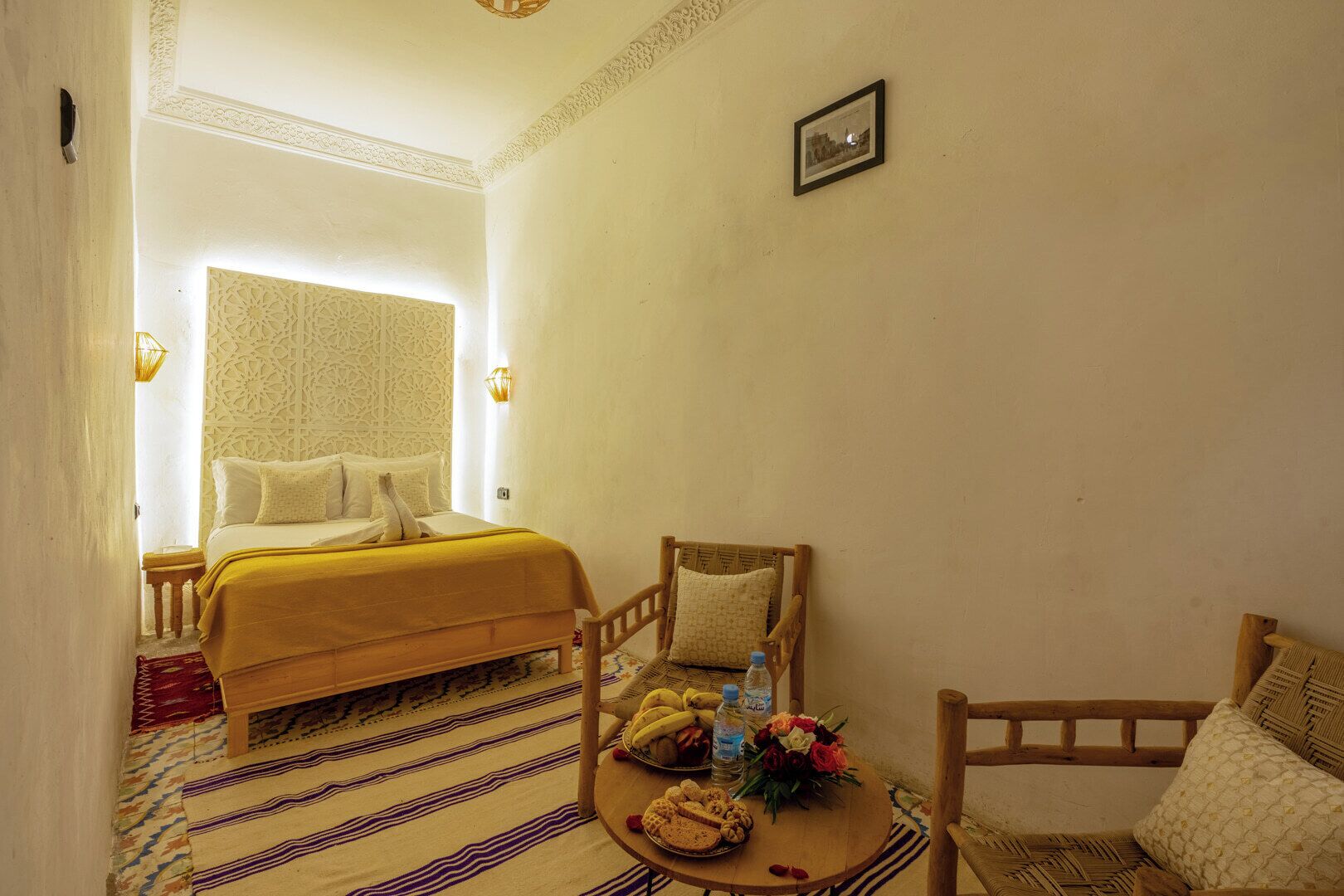 A room with a bed, a chair, a table with food and drinks, and a wall-mounted picture.