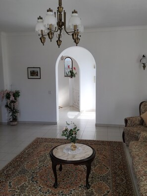 Uma sala de estar com um candelabro, um tapete estampado, um sofá, uma mesa de centro e uma porta que dá para outro cômodo.
