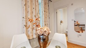 Un coin repas avec une table en verre, des chaises blanches, un vase métallique orné de fleurs et un rideau à motifs floraux.