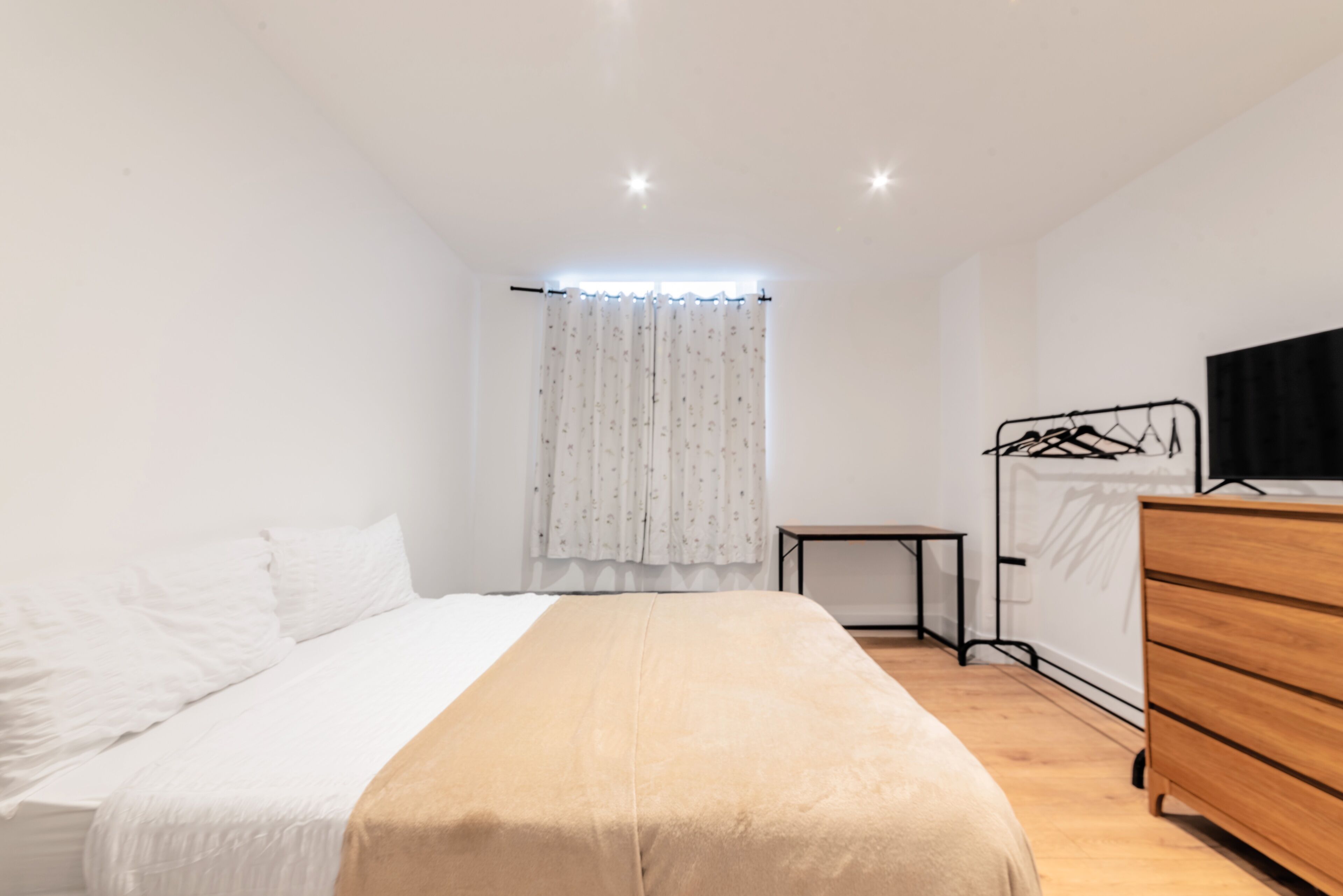 A bedroom with a bed, a dresser, a bedside table, a hanging clothes rack, and a window with curtains.