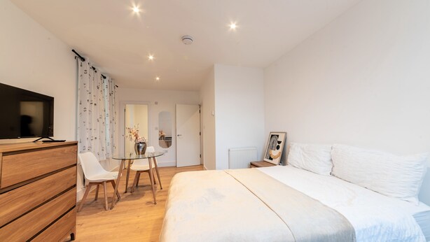 Une chambre à coucher comprenant un lit, une commode, un téléviseur, une petite table avec des chaises et une porte donnant sur une autre pièce.