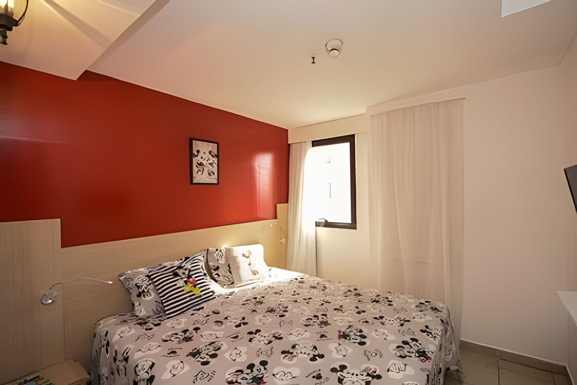 Un dormitorio con una pared de acento roja, una cama con cubrecama con temática de Mickey Mouse, una ventana con cortinas blancas y una lámpara de techo.