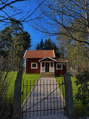 A red wooden house with a tiled roof, a front porch, and a gravel pathway leading to a gated entrance. - Cozy Cottage in top Condition Close to Nature and Lakes (Jonkoping)